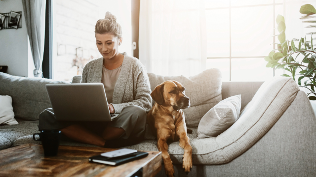 A woman sits cross-legged on a sofa, with her laptop on her lap. A brown dog sits next to her.
