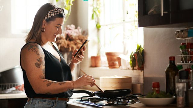A woman stirs some food in a pan, while smiling at her phone.