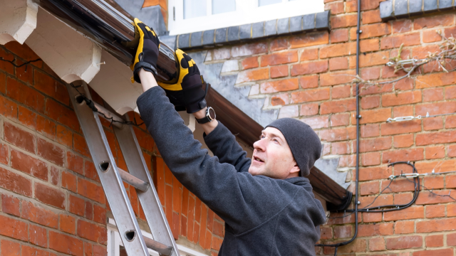 A man stands on a ladder, reaching up to a house's gutter.
