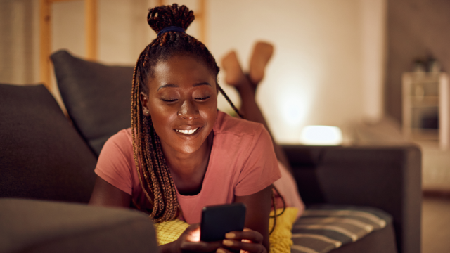 A woman lies on a sofa, smiling at her phone.