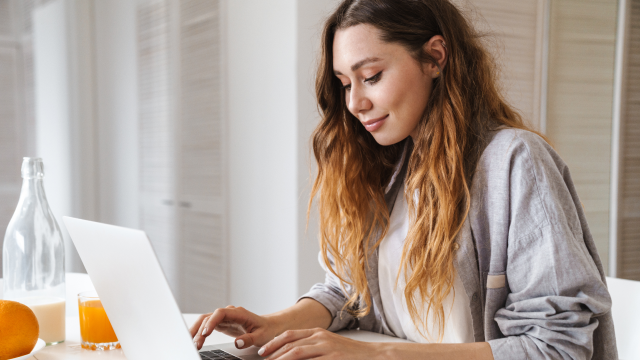 A woman with long sits working on her laptop, with a glass of orange juice beside her.