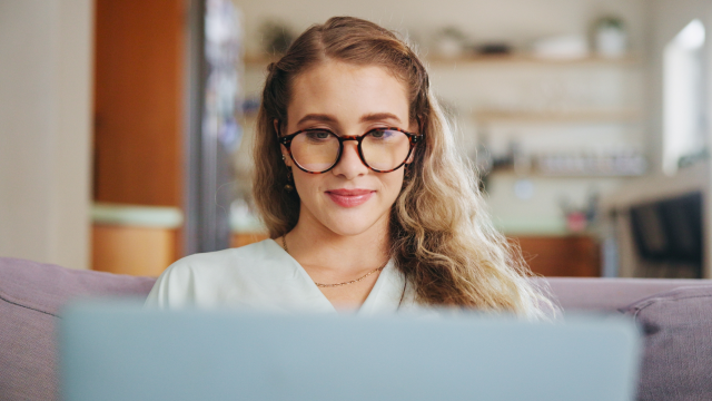 A woman with glasses sits on a sofa, typing on her laptop.