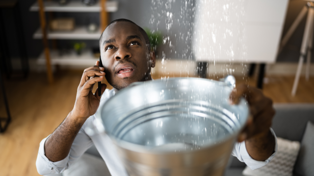 A man uses a bucket to catch a ceiling leak, while making a phone call.