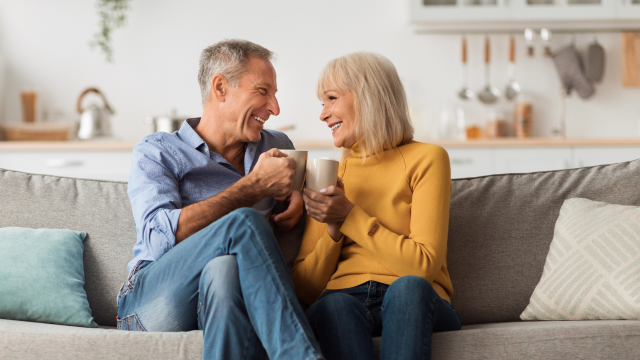 A couple of retirement age sit closely on the sofa, smiling at each other and while holding mugs.