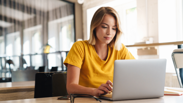 A woman in a yellow t-shirt sits at a desk, working on her laptop.