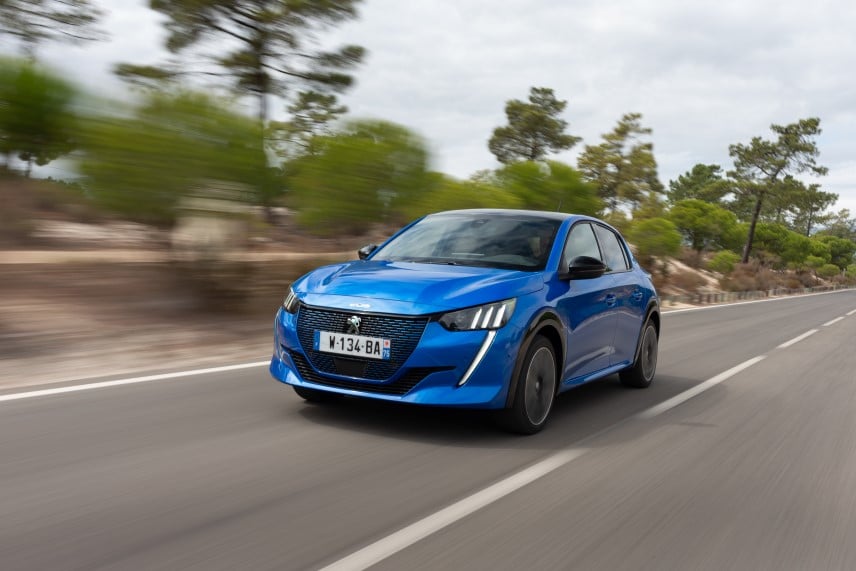 An image of a blue Peugeot e-208 driving along a road hedged by trees