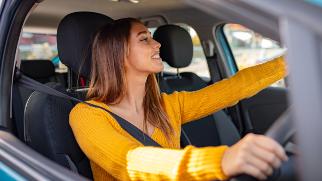 A woman sits in the driving seat of a car, with one hand on the wheel and one hand adjusting the rear-view mirror.