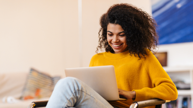 A woman in a yellow jumper sits on a chair, typing on her laptop and smiling.