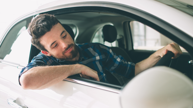 A man sits in the driver's seat of a car, with the window rolled down and his arm resting on the door.