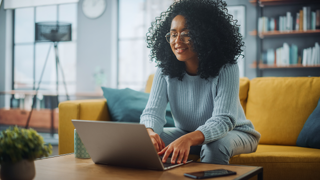 A woman sits on a sofa, typing on her laptop.