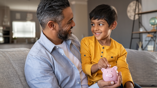A man and his young son sit on the sofa smiling at each other, as the son puts money into a piggy bank.