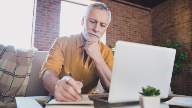 A man sits with his laptop open in front of him, writing in a notebook.