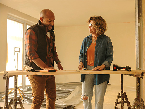 A man and a woman working on a home improvement project. They're in a room with protective coverings on the floor, standing behind a plank of wood resting on two sawhorses. Tools are placed on the plank of wood.