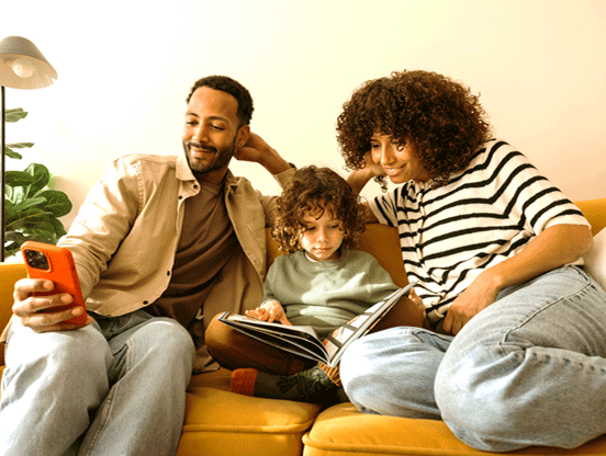 A young boy sits on the sofa reading a book with his mother. His father sits next to them, smiling at his phone.