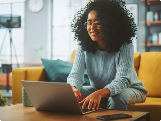 A woman sits on a yellow sofa, typing on her laptop